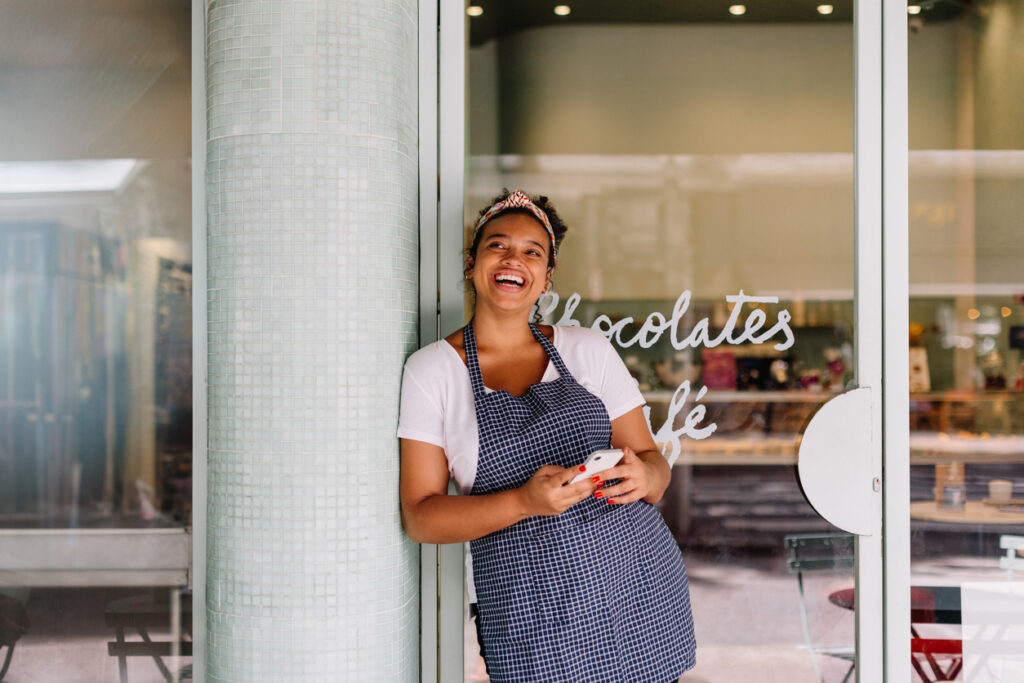 Successful cafe owner, a young woman, stands in her restaurant using a smartphone. Happy female entrepreneur using technology to manage her small business efficiently.