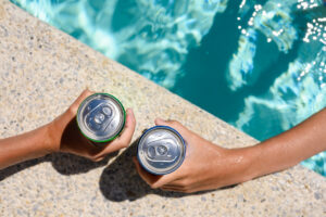 Two hands hold cans of cold drink in the pool on a hot day. The concept of vacation in a friendly company. Top view, selective focus on the bank.